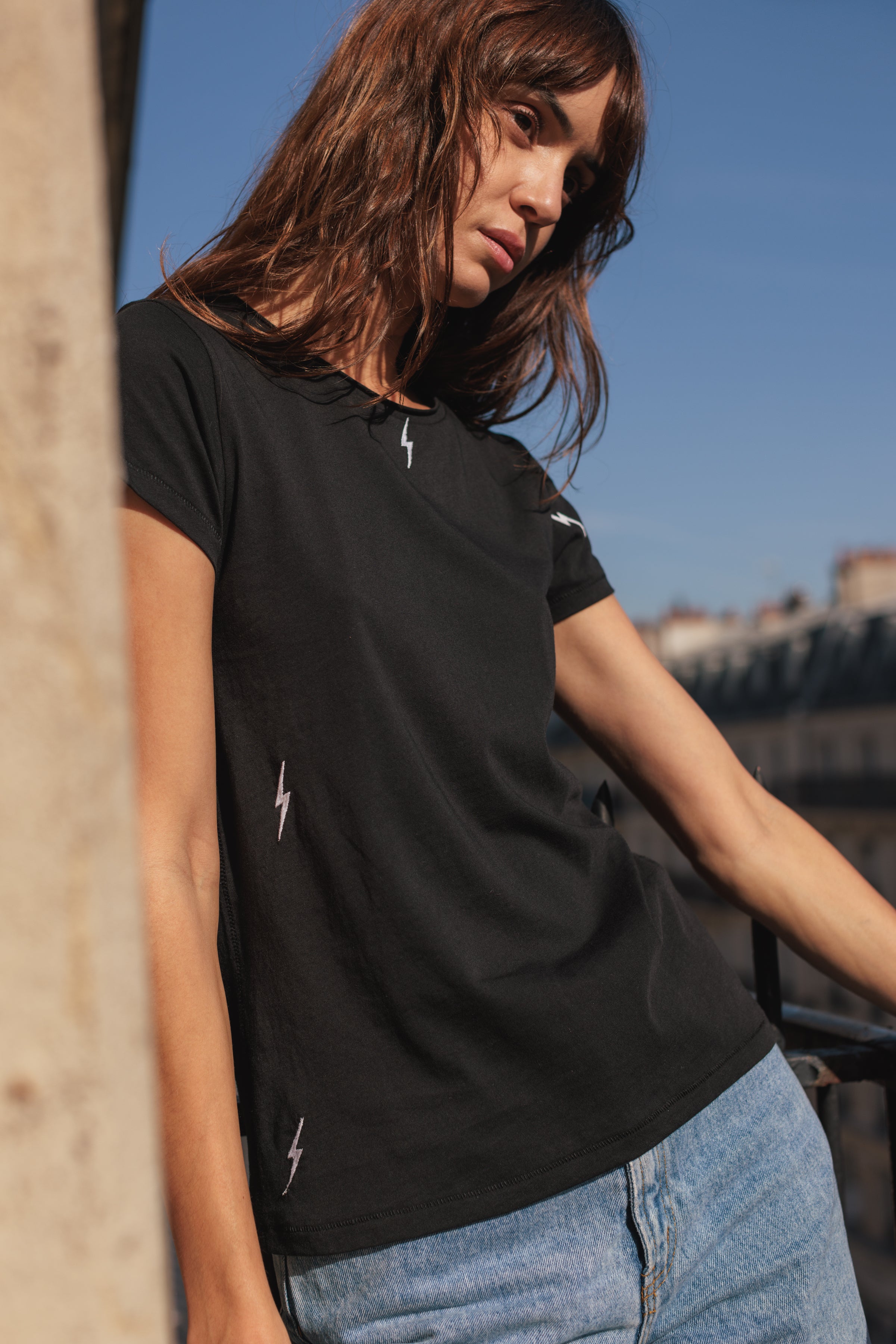 A woman wearing a Catherine Gee Embroidered Cotton Bolt black t-shirt with small, white lightning bolt embroidery scattered across the shirt paired with light blue jeans. The background features an outdoor setting with a clear blue sky and some buildings in the distance highlighting the casual yet stylish design of the t-shirt, making it interesting for fashion enthusiasts.