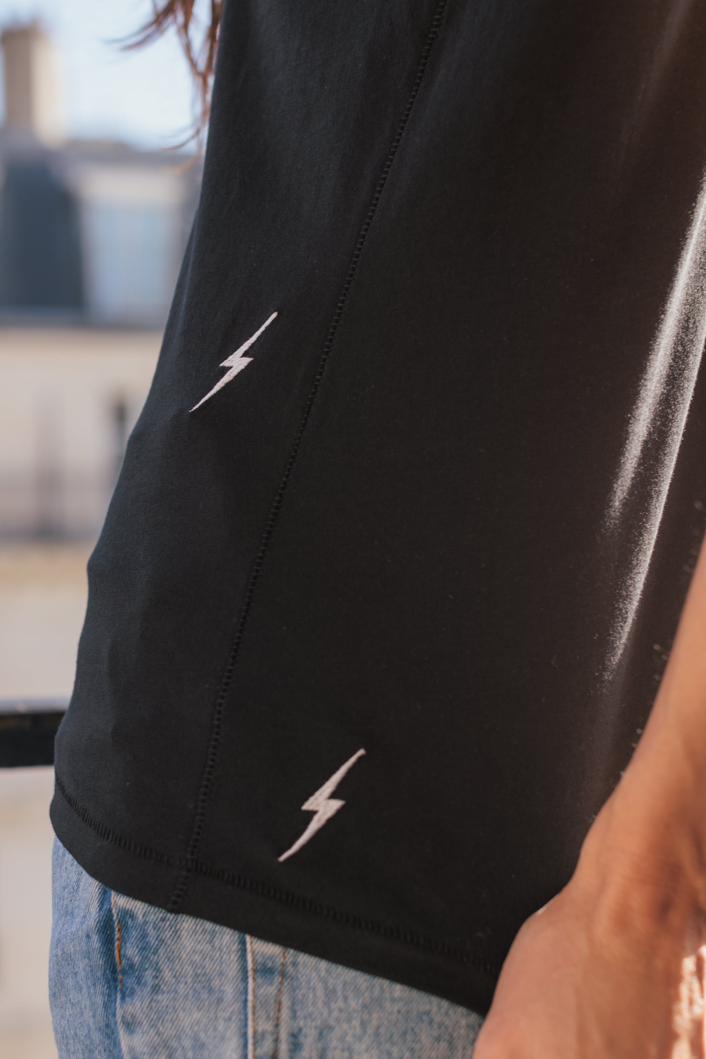 A close-up of a woman wearing a black color Catherine Gee Embroidered Cotton Bolt t-shirt features small, white lightning bolt embroidery designs, one near the side seam and the other near the bottom hem. She also wears light blue jeans highlighting the detailed embroidery and the casual yet stylish design of the t-shirt.