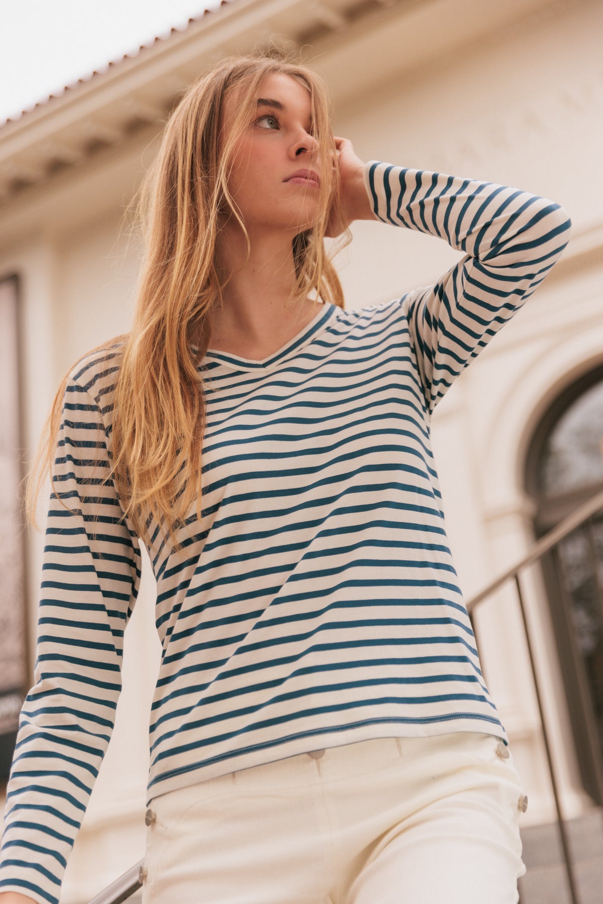 A woman wearing the Catherine Gee Jen Relaxed Longsleeve - Navy Stripe tee features classic navy and white horizontal stripes, a V-neckline, and long sleeves paired with light-colored pants. The background includes architectural elements, such as a building with arched windows and a staircase, adding a sophisticated urban feel to the image.