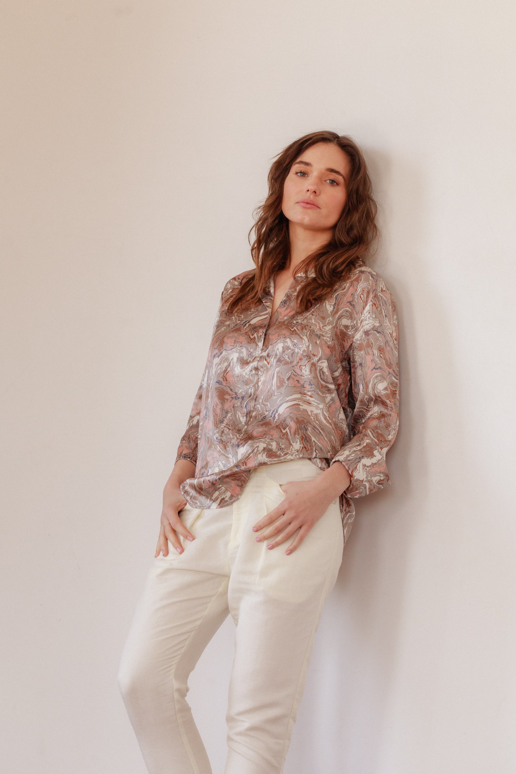 A woman standing against a plain background, with hands resting on the pockets of the pants wearing a Catherine Gee Daria French Cuff Silk Paris Paint blouse that features a detailed, abstract pattern with a mix of colors that resemble a painted design. The blouse has a loose fit and is paired with light-colored pants. The overall look is casual yet elegant, highlighting the unique design of the blouse.