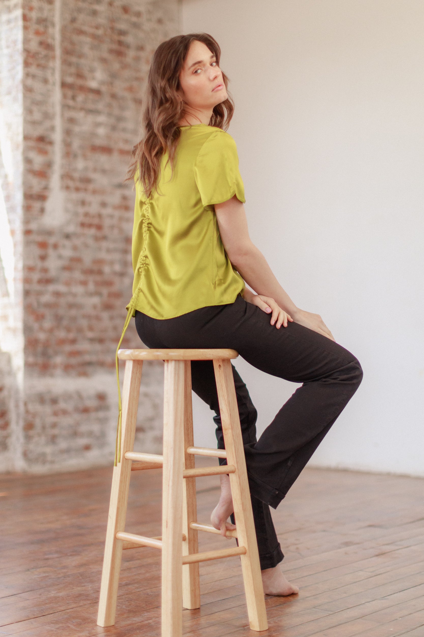 A woman is sitting on a wooden stool in a room with a brick wall and wooden floor wearing a bright green Catherine Gee Cate Ruched Back Shirt and black pants. The shirt features a distinctive ruched design on the back. 