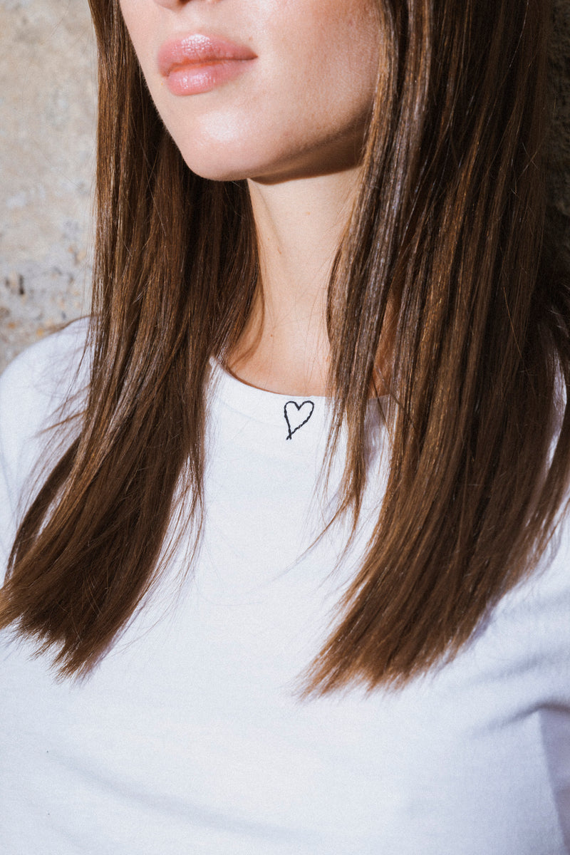 A close-up of a woman with straight, brown hair partially covering the white Catherine Gee Embroidered Cotton Heart t-shirt she wears, featuring a small, simple heart embroidered in a black thread near the neckline highlighting the minimalist and elegant design of the t-shirt, making it interesting for those looking for subtle yet stylish clothing options.