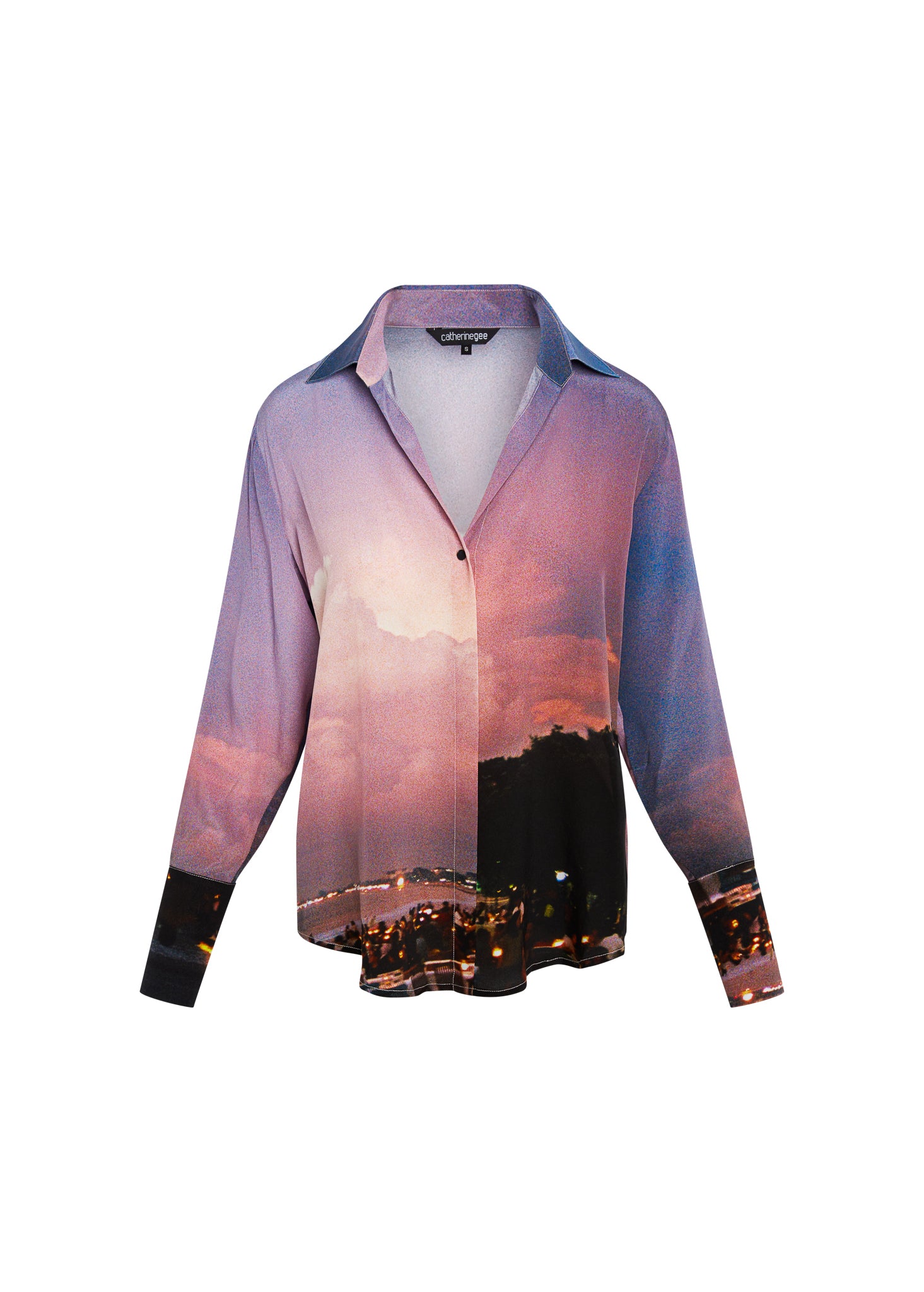 Catherine Gee Daria French Cuff Silk Bali Blouse features a vibrant print of a sunset over a cityscape, blending hues of pink, purple, and blue with city lights at the bottom. It has a classic collar, button-down front, and long sleeves with French cuffs.