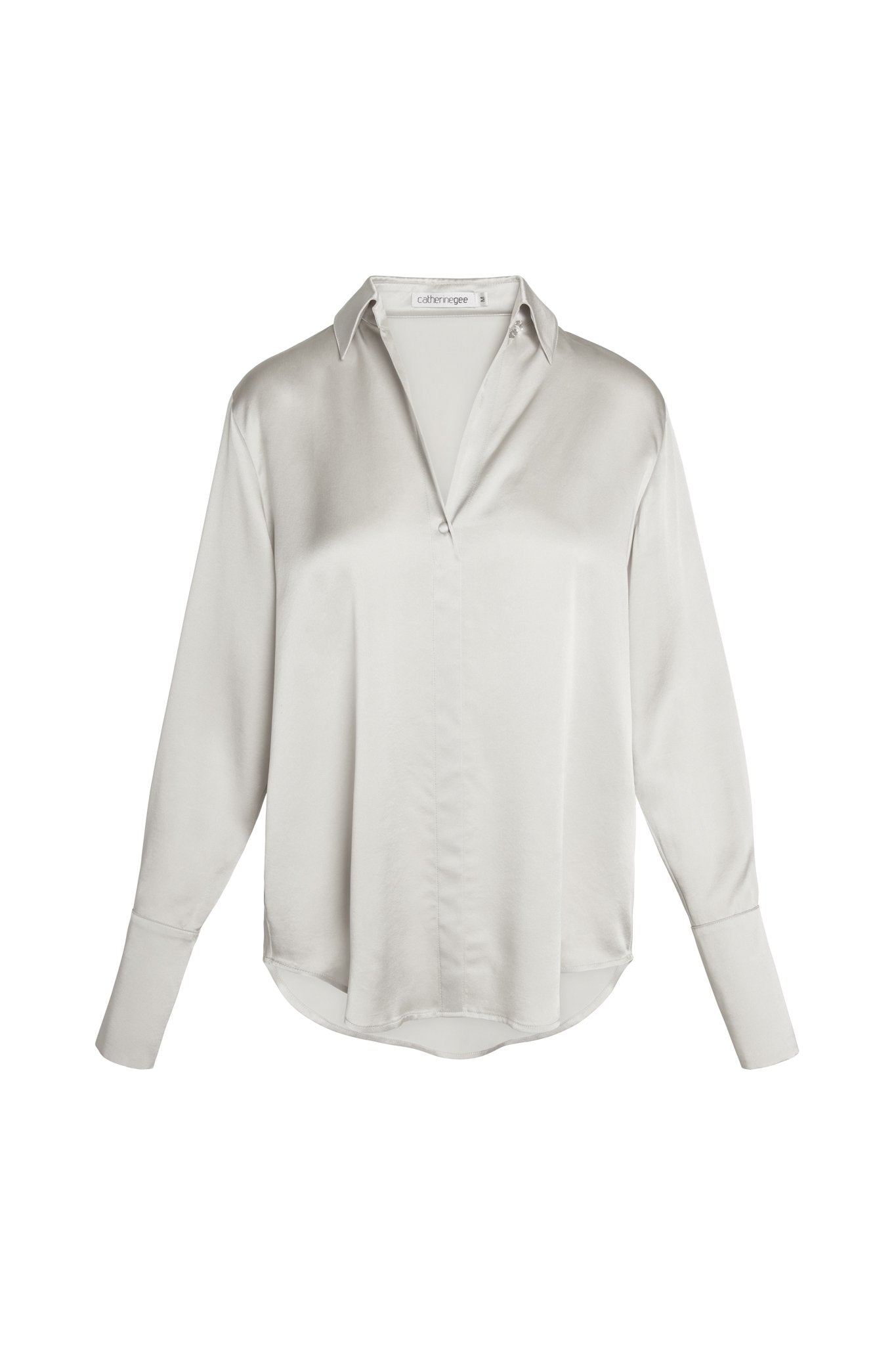 Catherine Gee Daria French Cuff Silk Silver Blouse. This elegant blouse is made from a smooth, shiny silk fabric in a silver color. It features a classic collar, a deep V-neckline, and long sleeves with French cuffs and has a relaxed fit with a slightly curved hemline, making it a sophisticated and versatile piece suitable for both professional and casual settings.
