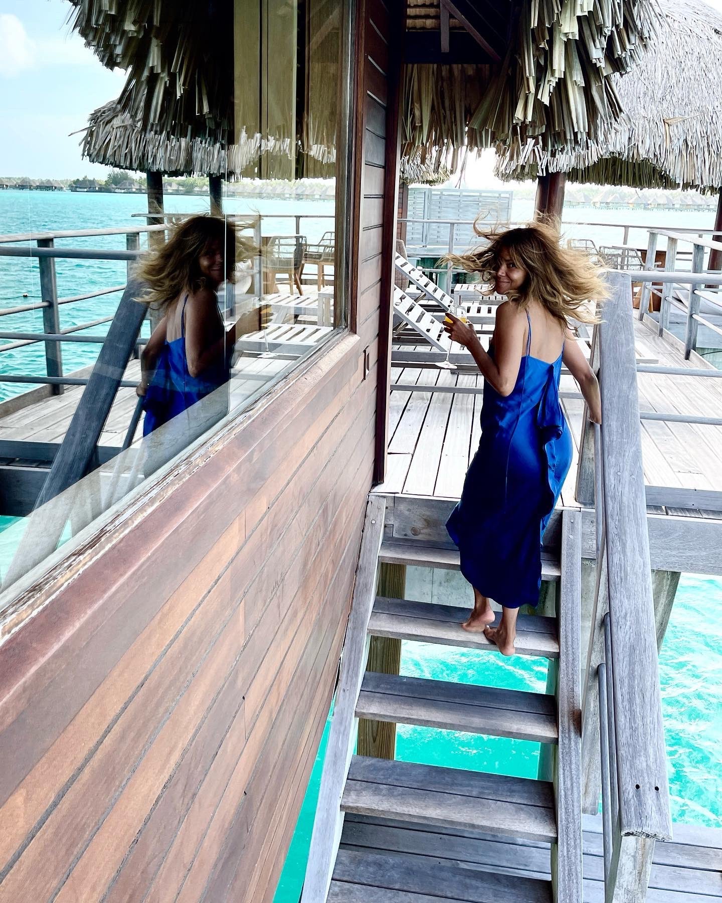 A woman wearing a Catherine Gee Emma Mykonos Blue Slip Dress, standing on a wooden staircase leading to an overwater bungalow. The dress is a vibrant blue, flowing elegantly as the person ascends the stairs. The setting is tropical, with clear turquoise water visible below and a thatched roof above, suggesting a luxurious vacation spot. The reflection of her in the glass window adds depth to the scene, enhancing the overall aesthetic appeal.