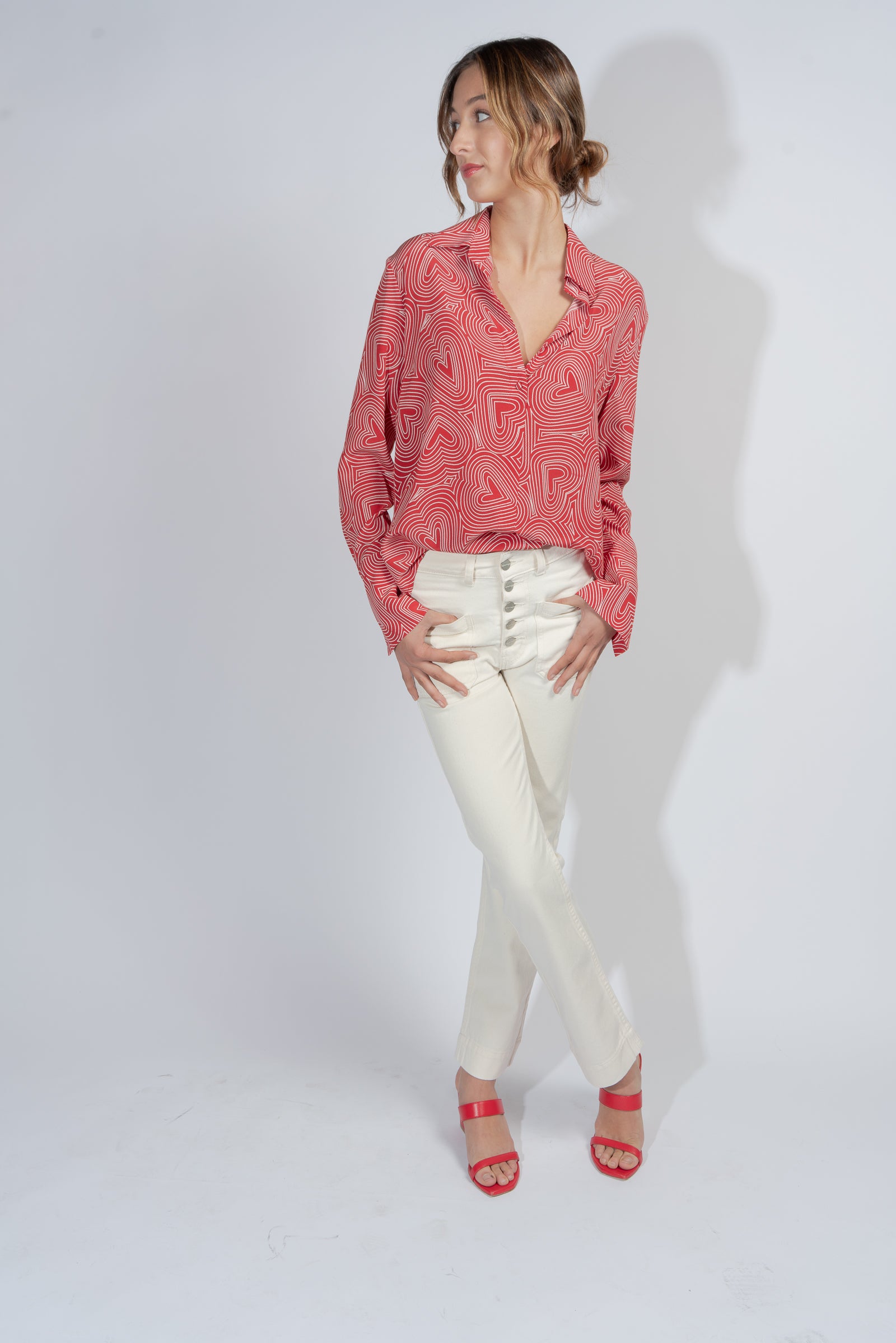 Woman wearing a Catherine Gee Daria French Cuff Silk Blouse - Bowie Heart. The blouse is red with white heart patterns and features long sleeves with French cuffs paired with white pants and red sandals, standing against a plain white background with legs crossed and hands resting on the thighs.