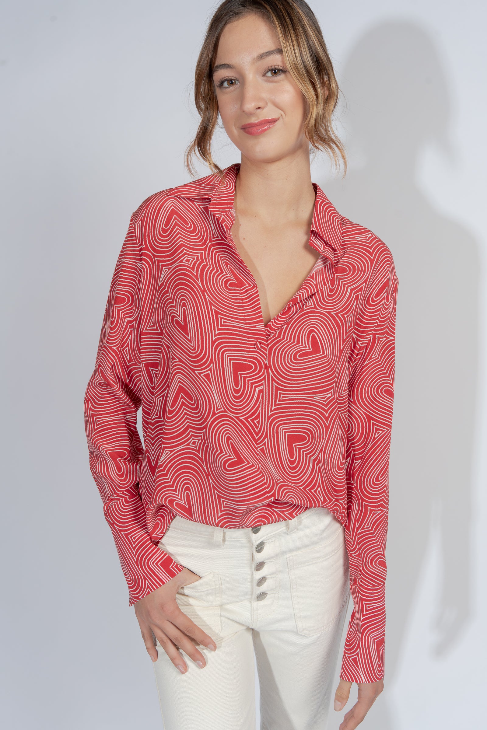 Woman wearing a Catherine Gee Daria French Cuff Silk Blouse - Bowie Heart. The blouse is red with a white heart pattern, featuring a deep V-neck and long sleeves paired with high-waisted white pants with multiple buttons.