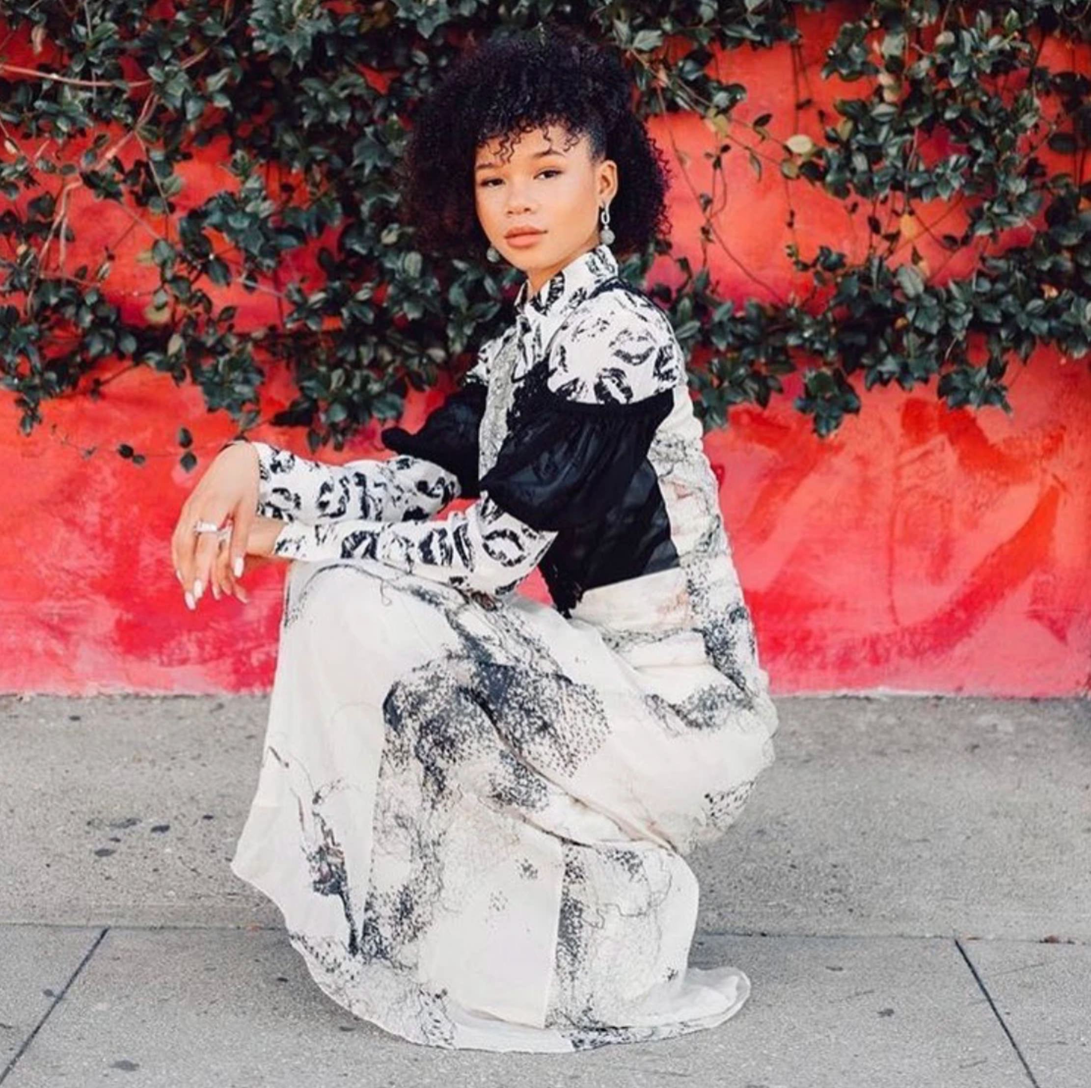 A woman is crouching in front of a vibrant red wall adorned with green foliage. She is wearing a Catherine Gee Daria French Cuff Silk Blouse with a black and white lip print design that features long sleeves and a high collar, paired with a matching skirt that has a similar black and white print. The outfit stands out against the colorful background, creating a striking visual contrast.