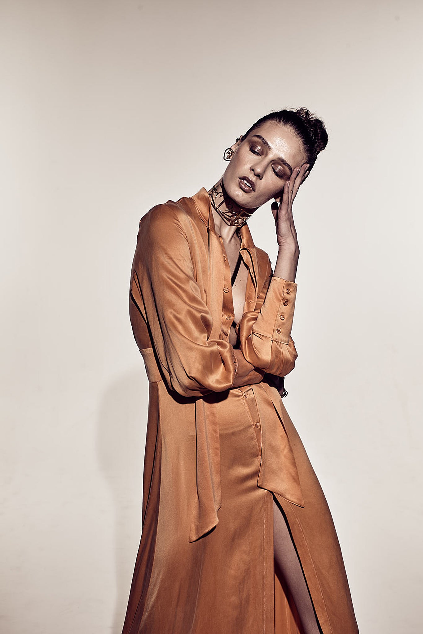 A woman posing with one hand touching the face wearing a Catherine Gee Electric Flou High-Slit Long dress is a long, flowing garment with a high slit on one side, allowing for a glimpse of the leg. It features a button-down front and long sleeves, with a loose, elegant fit. The fabric appears to be silky and smooth, giving the dress a luxurious and sophisticated look adding a sense of style and grace to the overall appearance.