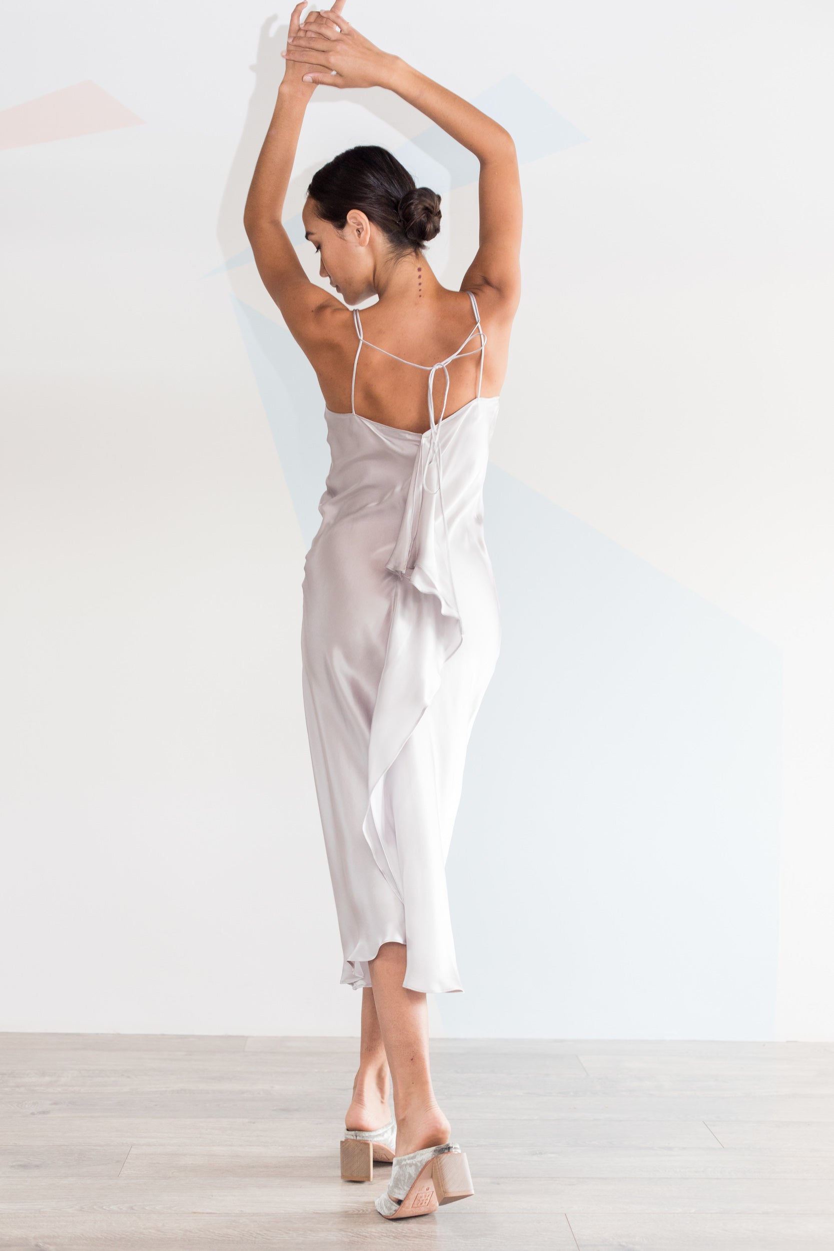 A woman wearing the Catherine Gee Emma Silver Slip Dress. This sleek and elegant dress is made of a shiny silver fabric with a smooth, flowing silhouette. It features thin spaghetti straps that crisscross at the back, creating a sophisticated look. The dress includes a ruffled detail cascading down the back, adding femininity and movement. She stands on a light wooden floor, arms raised, showcasing the dress's stunning back design.